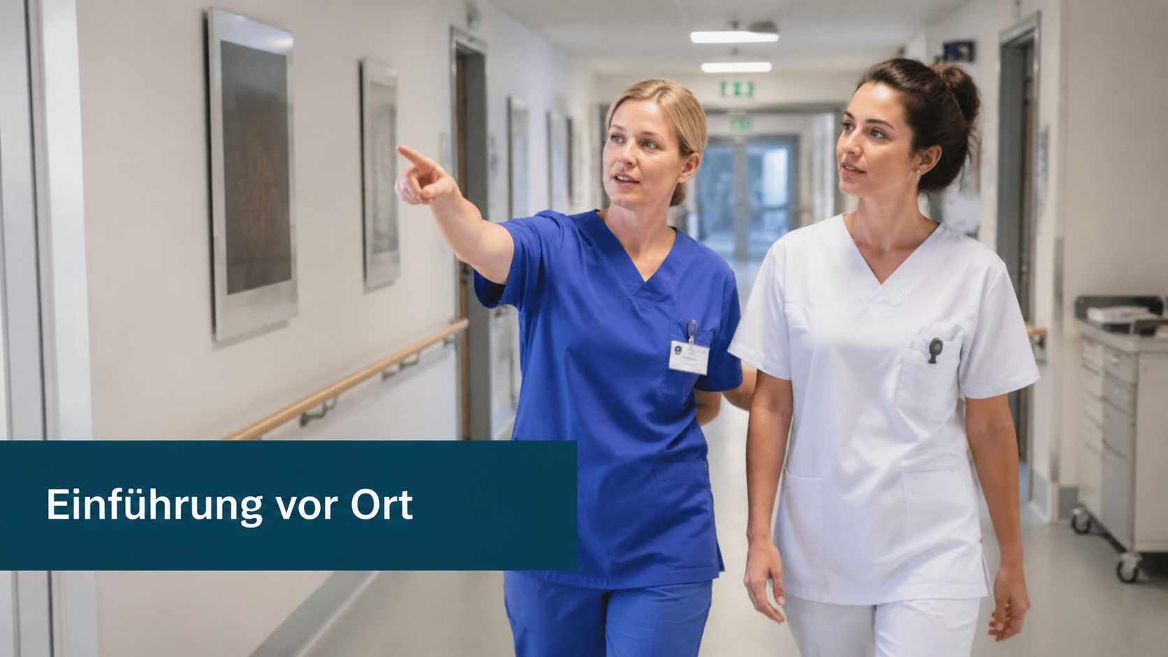 Eine Krankenschwester in blauer Uniform zeigt ihrer Kollegin in weißer Kleidung den Krankenhausflur während der Einarbeitung.